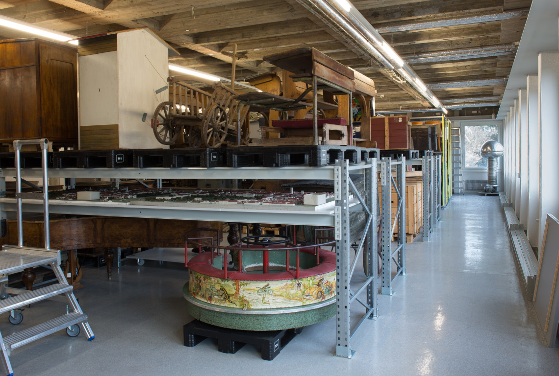Blick in ein Depot des Stadtmuseums. In langen Metallregalen stehen unter anderem alte Schulbänke, ein Leiterwagen und ein Kinderkarussell.